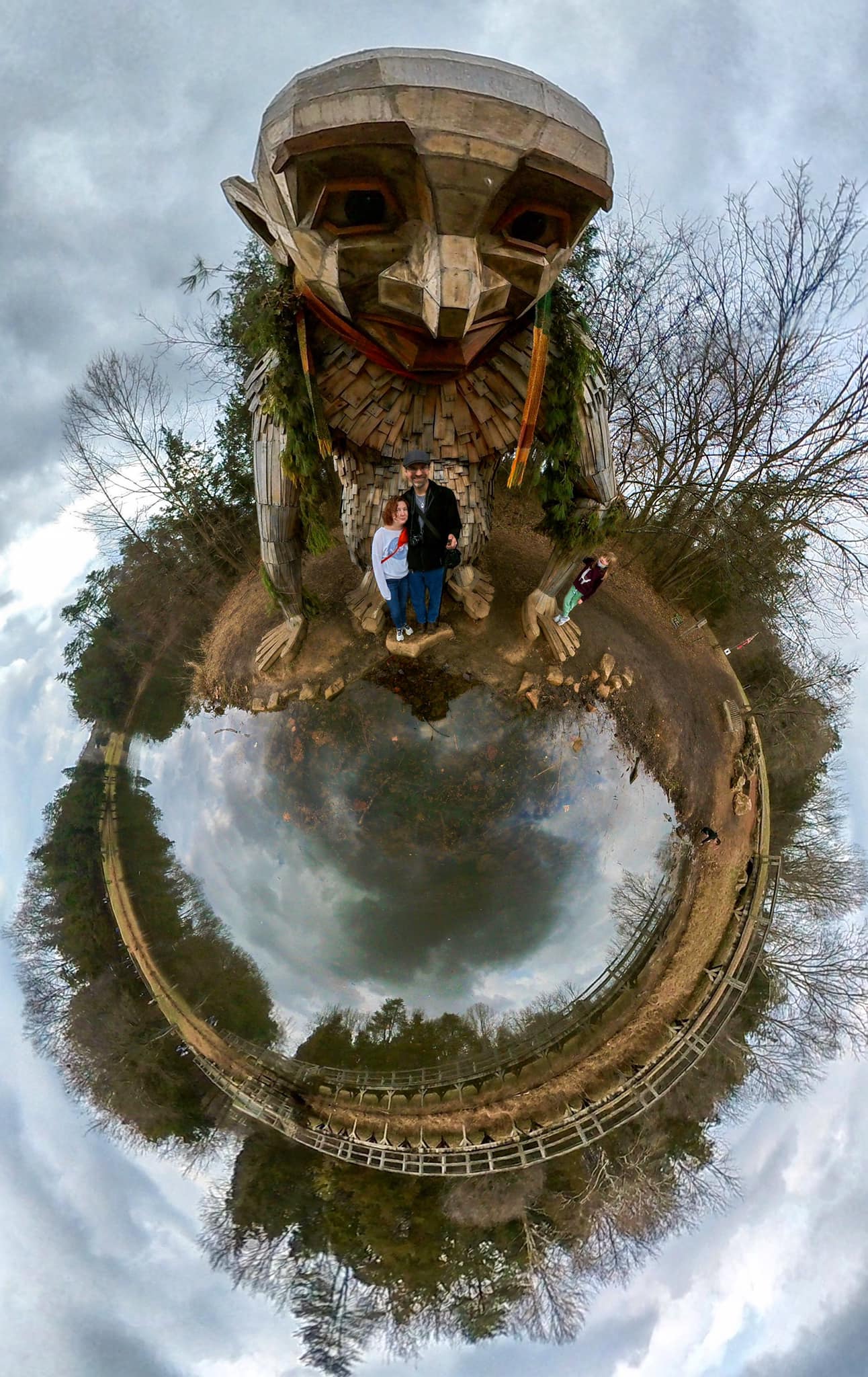 A tiny planet image of a large wooden troll at a reflecting pond with people beneath it