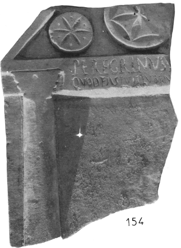 black and white photo of a broken altar with oone and a half sand dollars at the top, and an inscription reading Peregrinus quod Esus fuit iuben. There is a carved pillar but no other decoration.