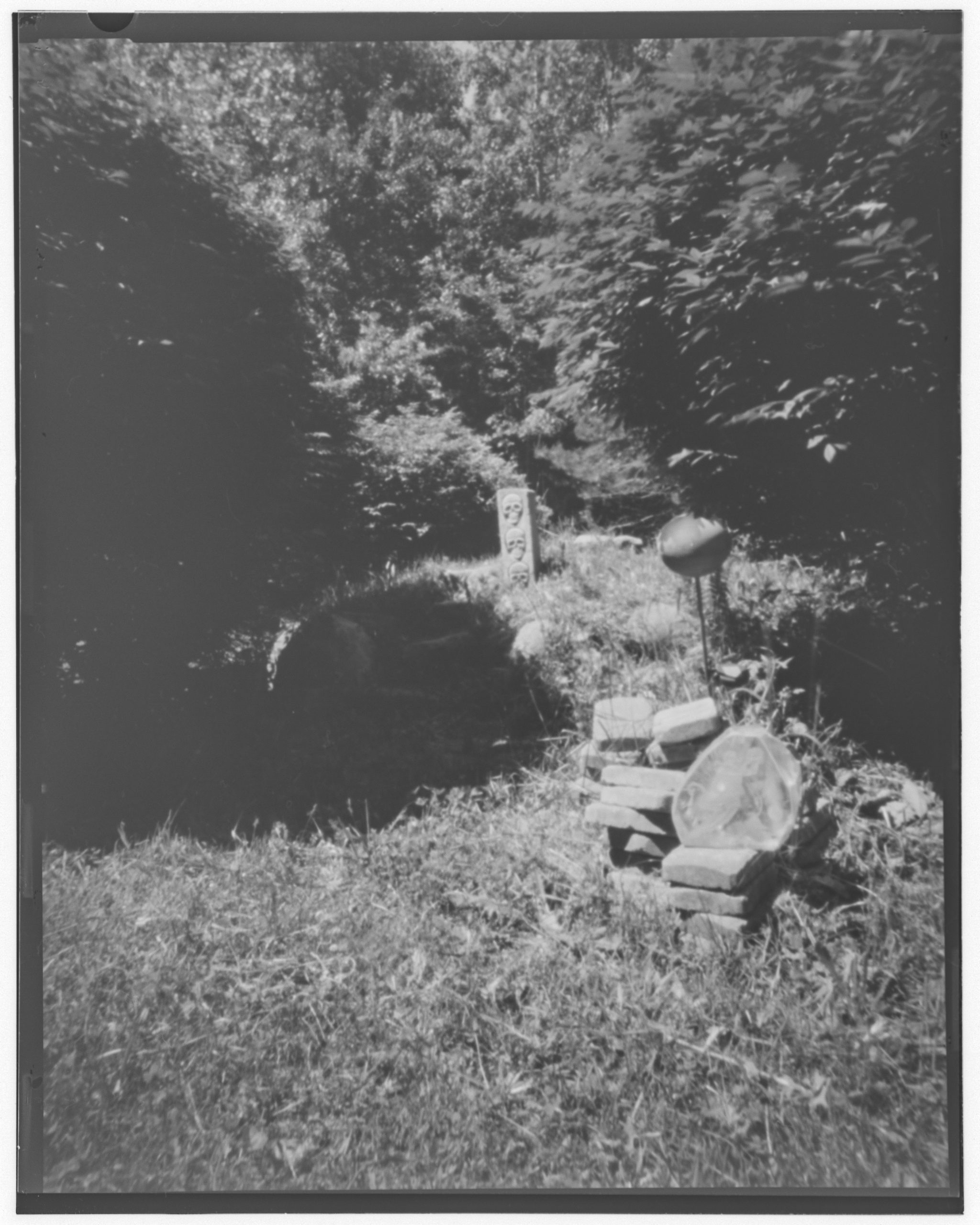 Pinhole black and white image of a mound of dirt, surrounded by trees, with a stone pillar with carved skulls on it