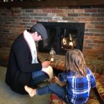 A child sits next to Rev. Dangler as he lights a candle from a lantern next to a lit fireplace