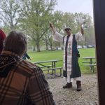 MJD stands in the rain in ritual garb, holding up his sickle and a pitcher of water