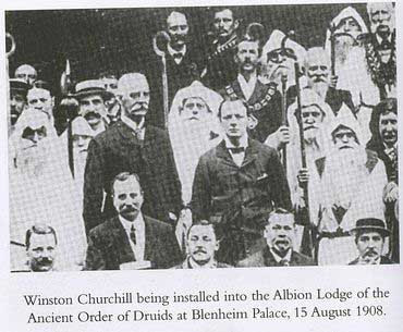Two men in suits surrounded by men in white robes and long fake white beards. Text: "Winston Churchill being installed in to the Albion Lodge of the Ancient Order of Druids at Blenheim Palace, 15 August 1908."
