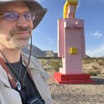 MJD takes a selfie with a large pink, yellow, and red cinderblock statue of a nude woman in the desert