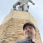 MJD under a statue of Vulcan, whose bare butt is turned to him, high above his head