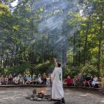 MJD in the center of a circle next to a fire, a hand raised. Summerland attendees on benches in the background