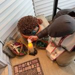 MJD lights a candle from another candle, kneeling on a front porch in the snow, in front of a corn dolly