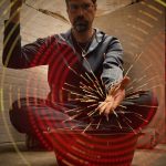 MJD sits with his right hand pointed up and left hand out; from his left hand postproduction sparks radiate