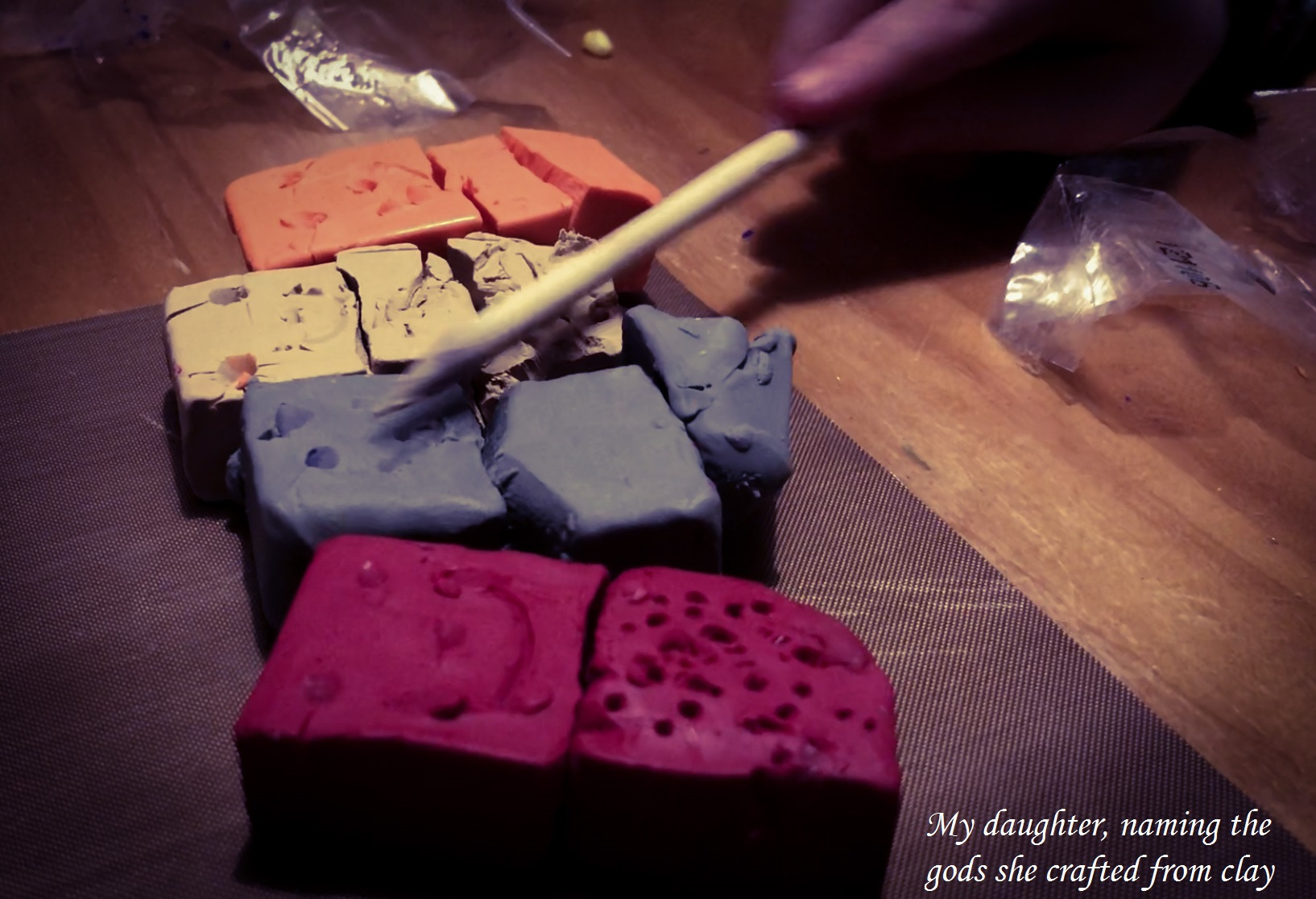 blocky, rough-shaped clay figures with smiley faces carved into them; text: "my daughter, naming the gods she crafted from clay"