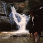 MJD wearing a hiking backpack stands before a waterfall